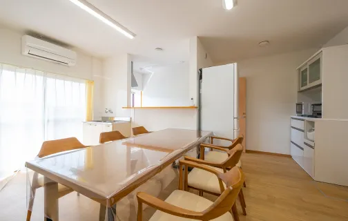 写真：7人掛けのダイニングテーブルがあり、対面式の台所や調理器具の置いてある棚があるダイニングキッチン。